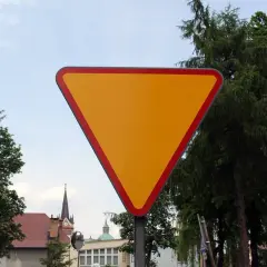 Żółty trójkątny znak drogowy z czerwoną obwódką, na tle drzew i nieba.