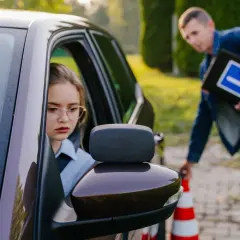 Dziewczyna w okularach za kierownicą samochodu, instruktor z tablicą "L" obok pachołka.