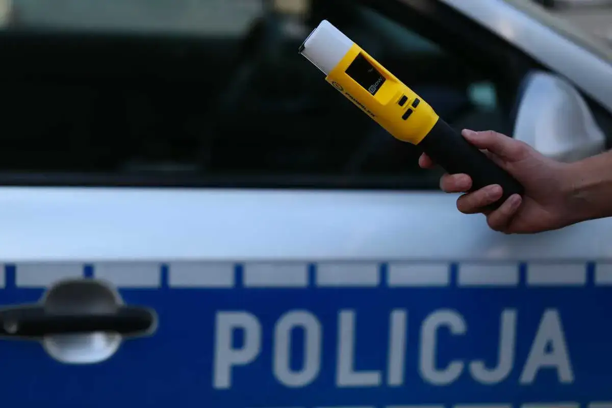 Dłoń trzymająca żółty alkomat na tle radiowozu z napisem "POLICJA".