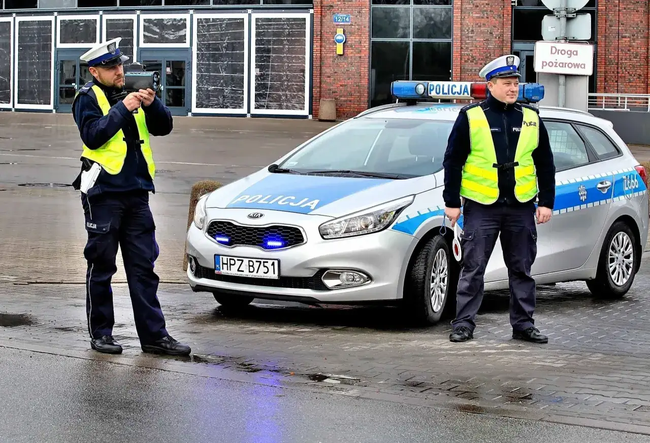 Policjanci z radarem mierzą prędkość. Jeden z nich celuje w kierunku drogi, drugi stoi obok radiowozu. Kierowcy, uważajcie na przekroczenie prędkości o 20 km/h!