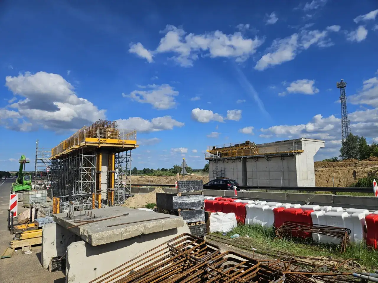 Budowa wiaduktu na autostradzie A6. Betonowe filary i rusztowania, w tle niebo z chmurami i wieża telekomunikacyjna.